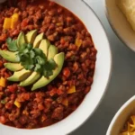 A bowl of Keto Taco Soup topped with avocado slices and cilantro, served alongside shredded cheese and crispy tortilla chips.