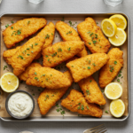 A tray of golden, crispy fish fillets made using the Hillbilly Fish Fry Seasonings Recipe, served with lemon wedges, tartar sauce, coleslaw, hushpuppies, and fries.